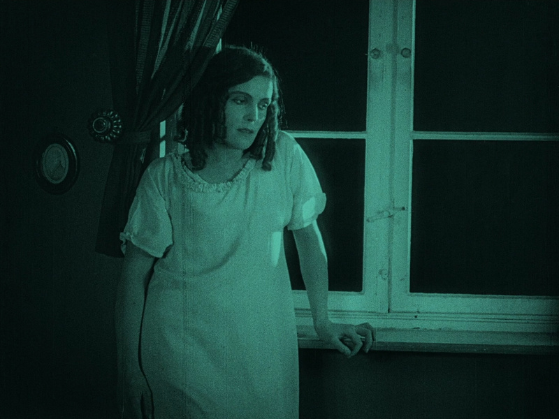 A famished woman standily wearily beside her bedroom window.