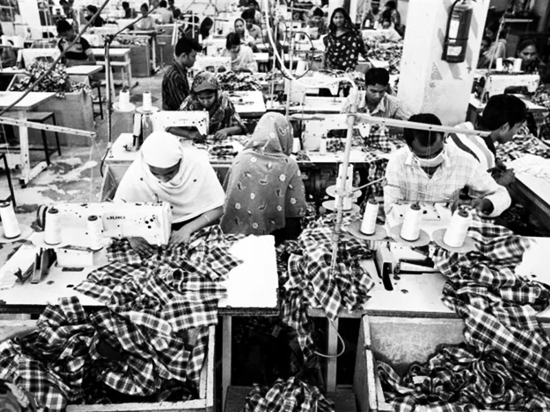 A dozen sweat shop employees work in cramped conditions. This image is in greyscale for dramatic effect.